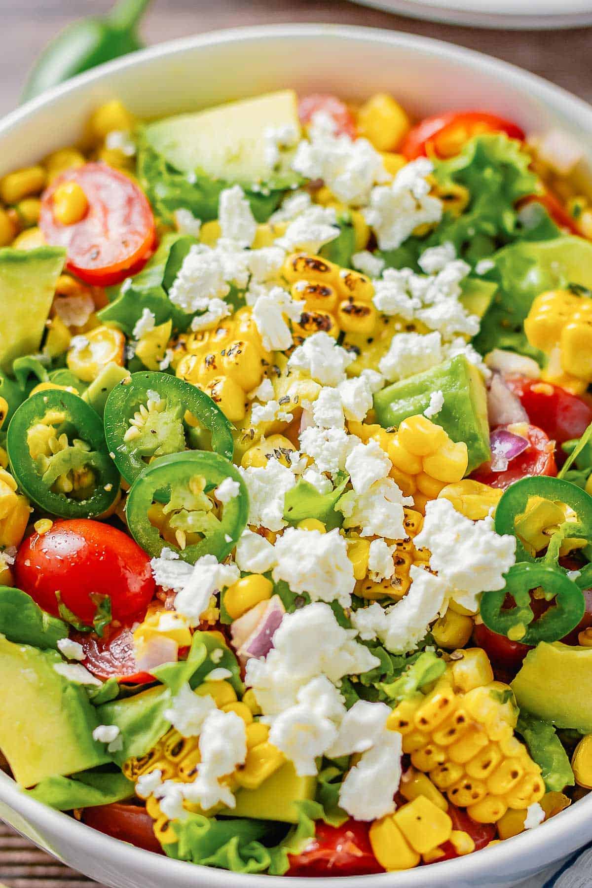 close up shot of a grilled corn salad with lettuce, avocado, tomatoes, onion in a white bowl on a wooden table