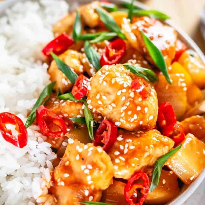 chicken chunks with bell peppers and pineapple in rich sweet and sour sauce in a bowl garnished with chili slices and scallion